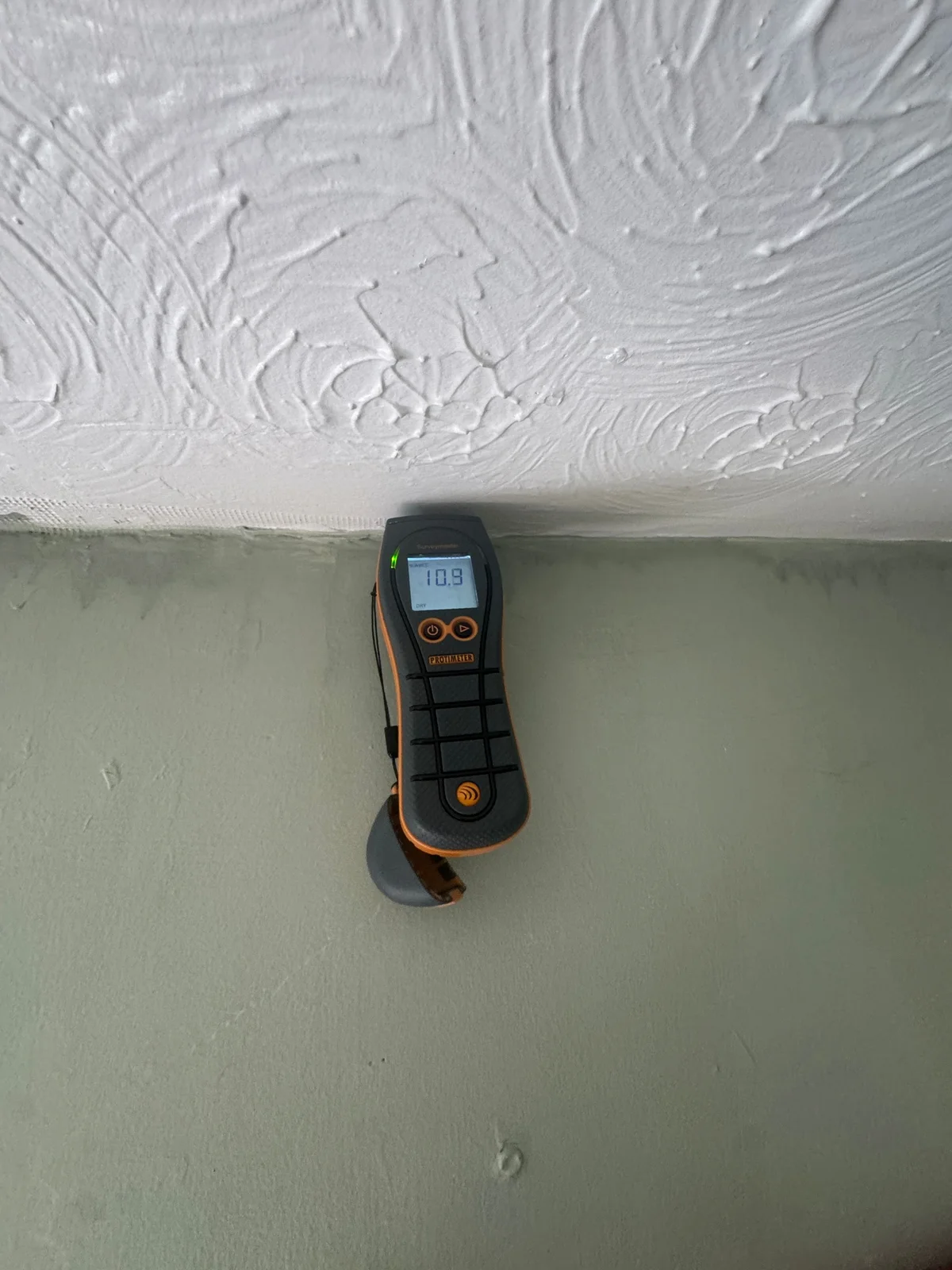 A moisture meter shows a dry indication beside visible marks at the top of a decorated wall.